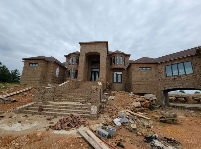 Large brick house under construction with stone steps and overcast sky.