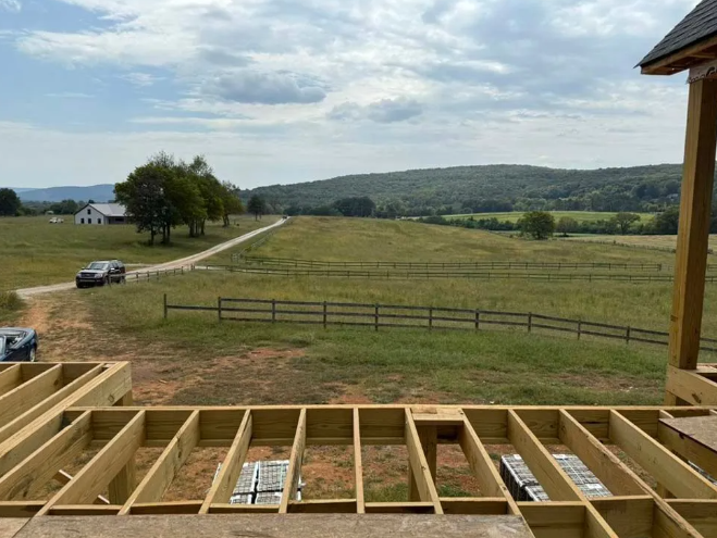 Wooden frame of a deck with a vast green field, road, and mountains in the distance under a cloudy sky.