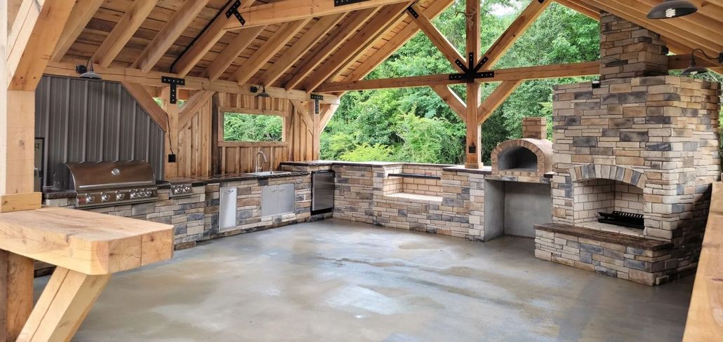 Outdoor kitchen with stone accents, a built-in grill, and a pizza oven under a wood-beamed roof.