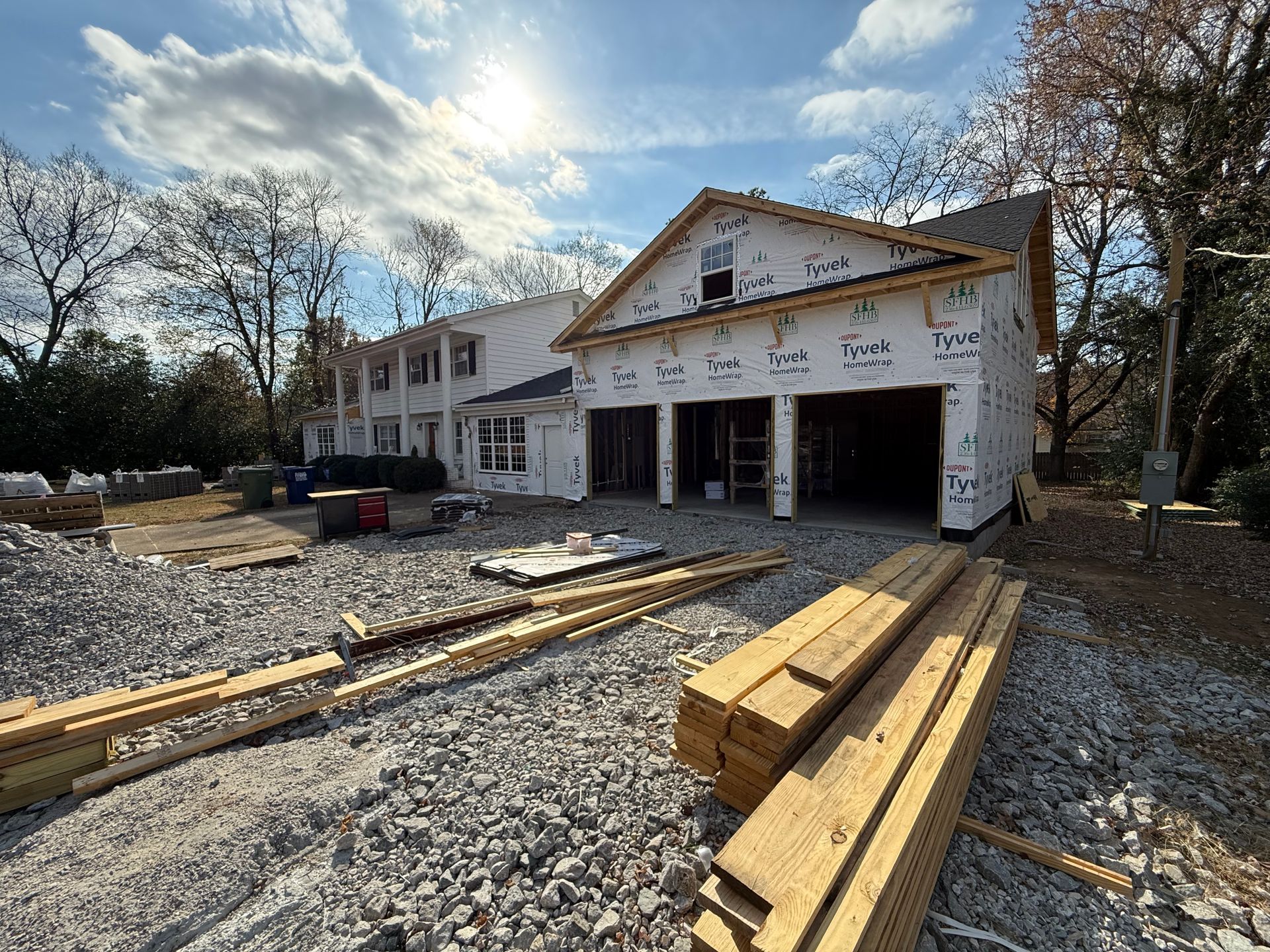 Construction site with a partially built house and garage. Lumber, gravel, and trees surround the structure under a blue sky.