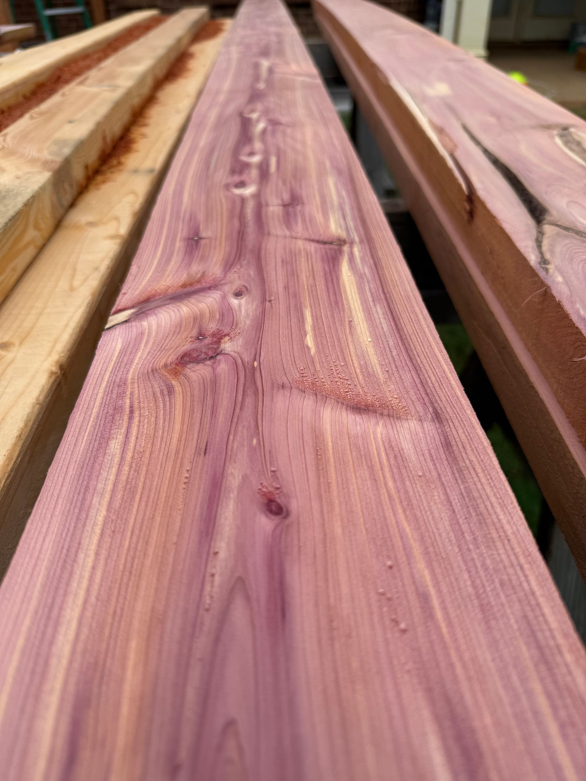 Close-up of a long, purple-hued cedar wood plank with visible grain and knots. Other planks in the background.
