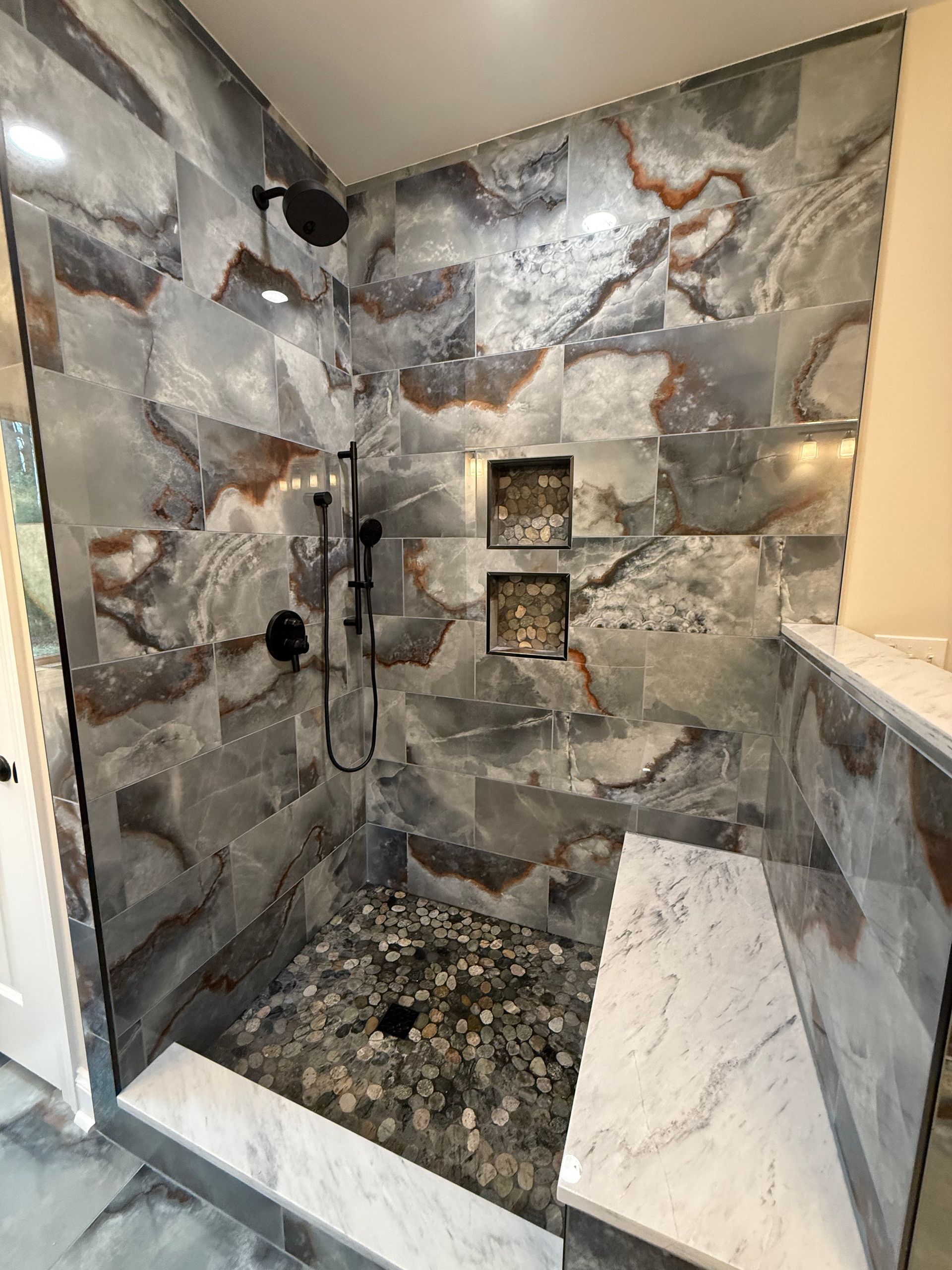 Stone-tiled walk-in shower with black fixtures, pebbled floor, and built-in niche shelves.