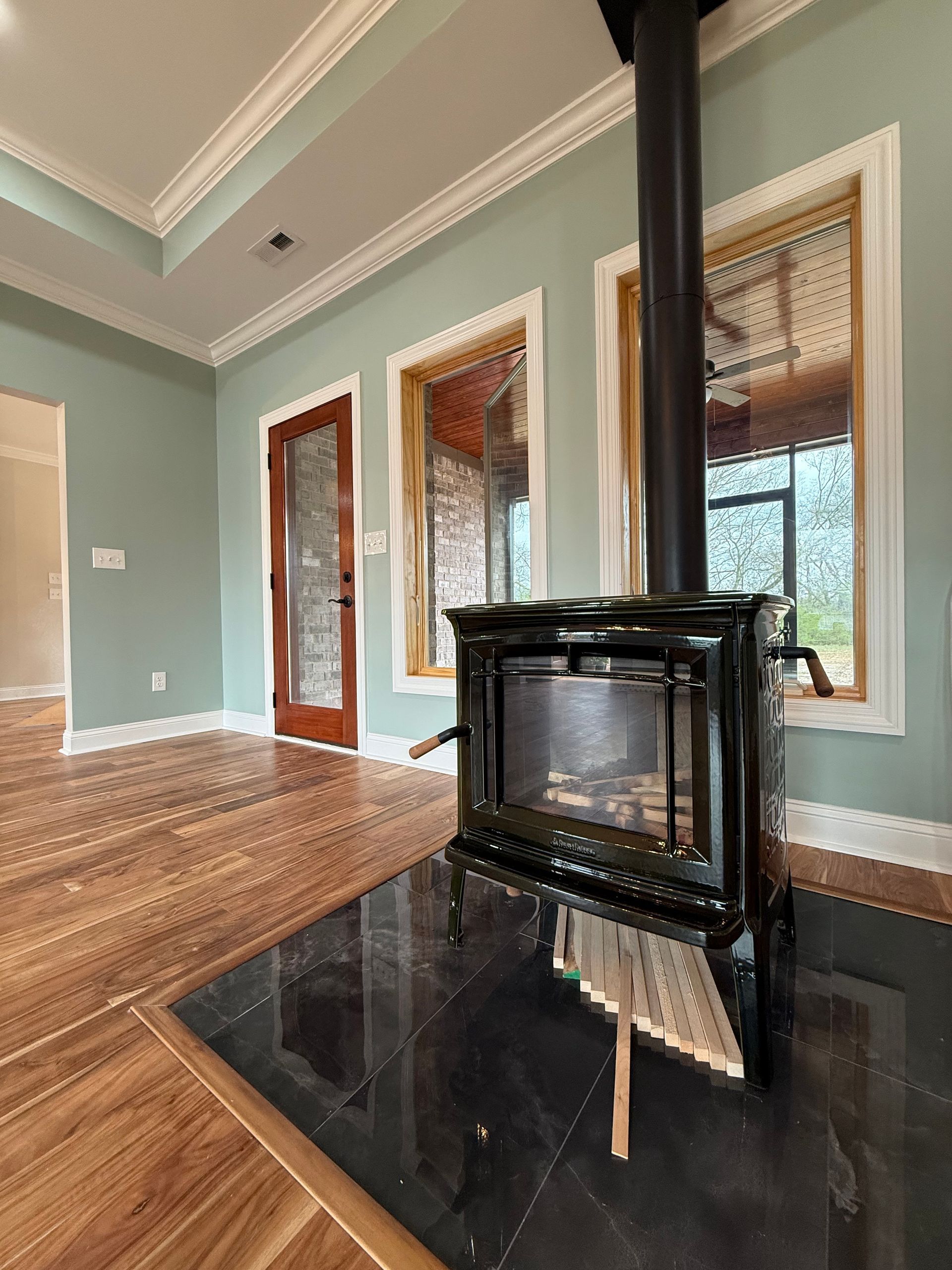 Modern living room with green walls, wood floors, a black stove on a hearth, and tall windows with blinds