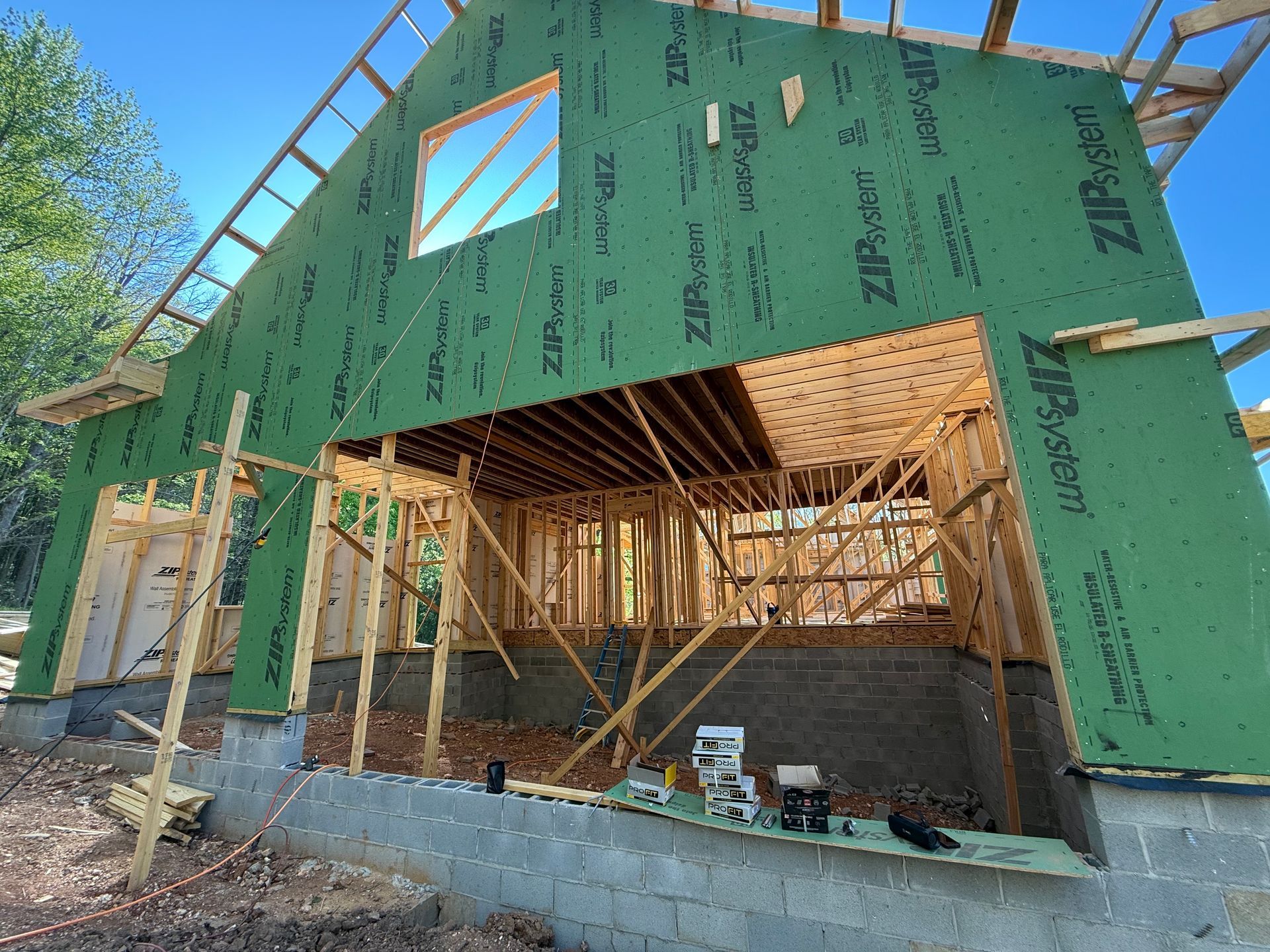 Partially built house framed in wood and wrapped in green housewrap, with an open front and stone foundation.
