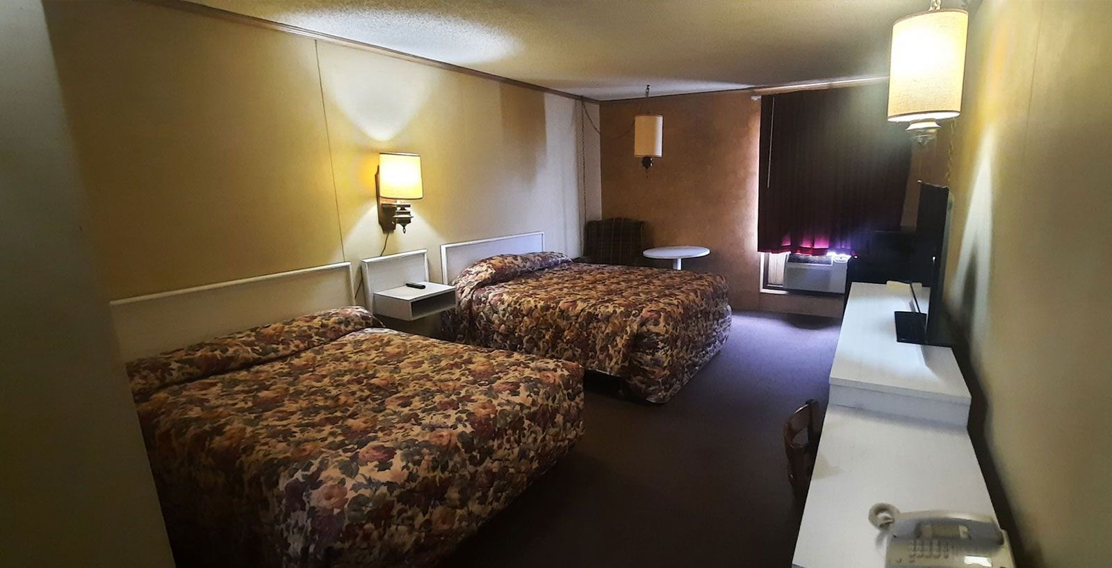 A motel room with two beds, 3 lamp shades, and a long desk with television and phone