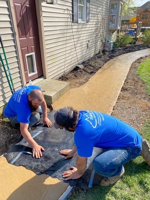 Two men in blue shirts are working on a sidewalk in front of a house.