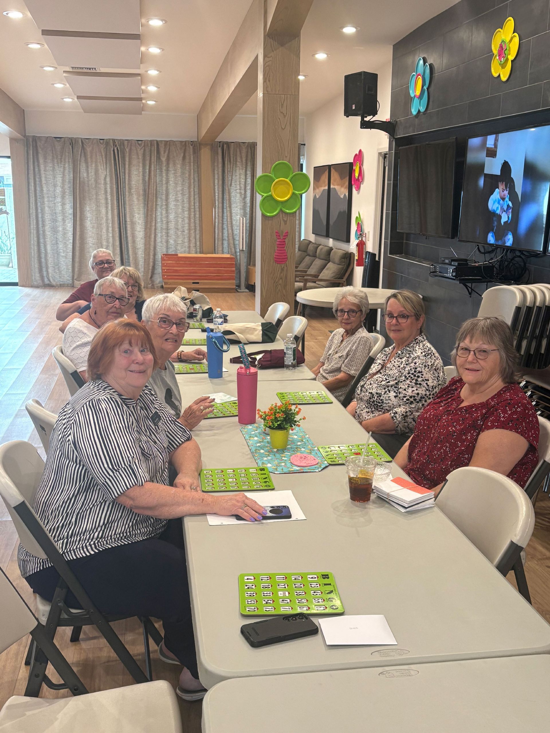 A group of senior women playing bingo at a long table in a community center. The setting includes decorations, a TV, and a lounge area.