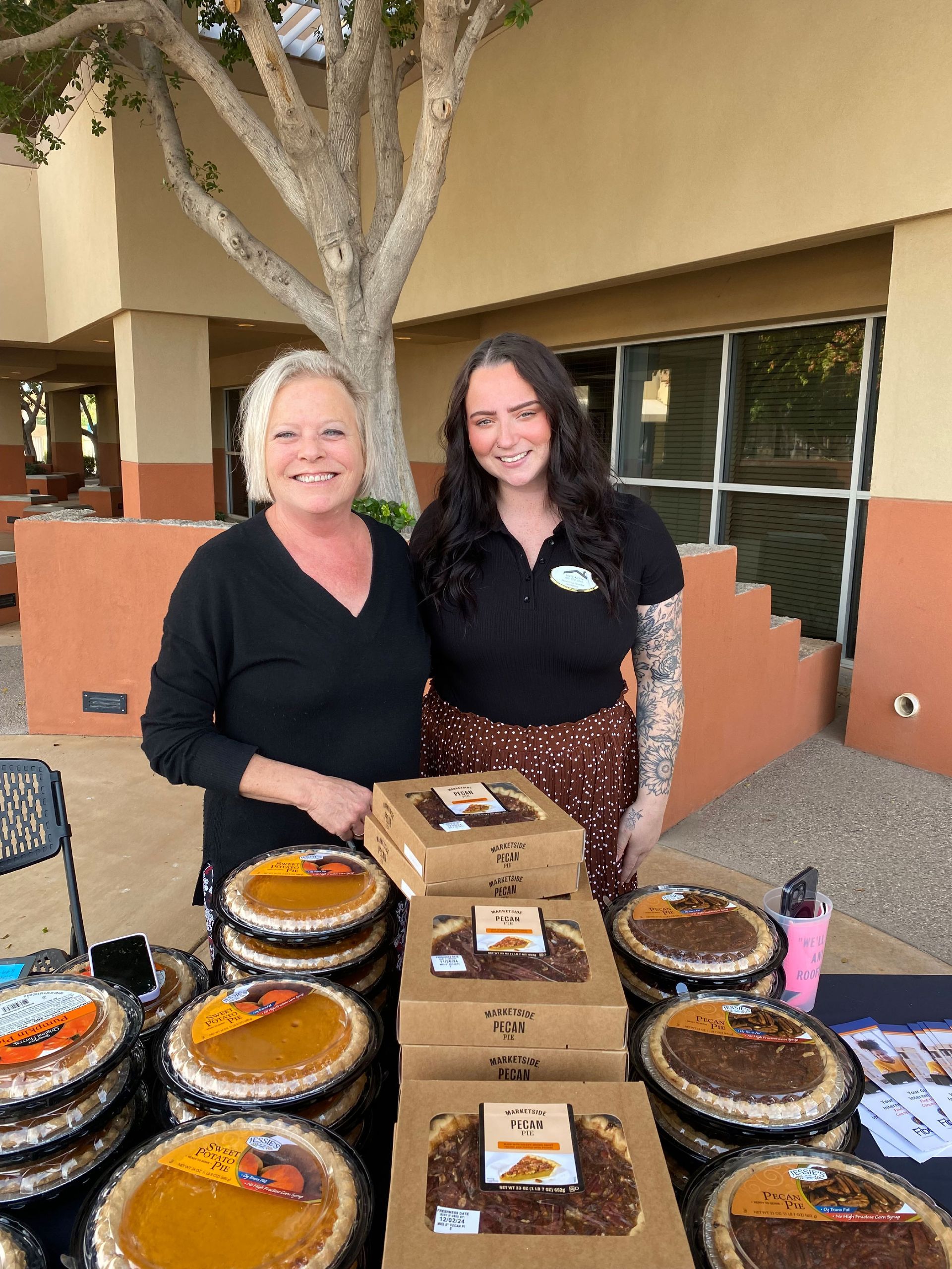 Two women stand behind a table selling pies outdoors. Pies are stacked in front of them.