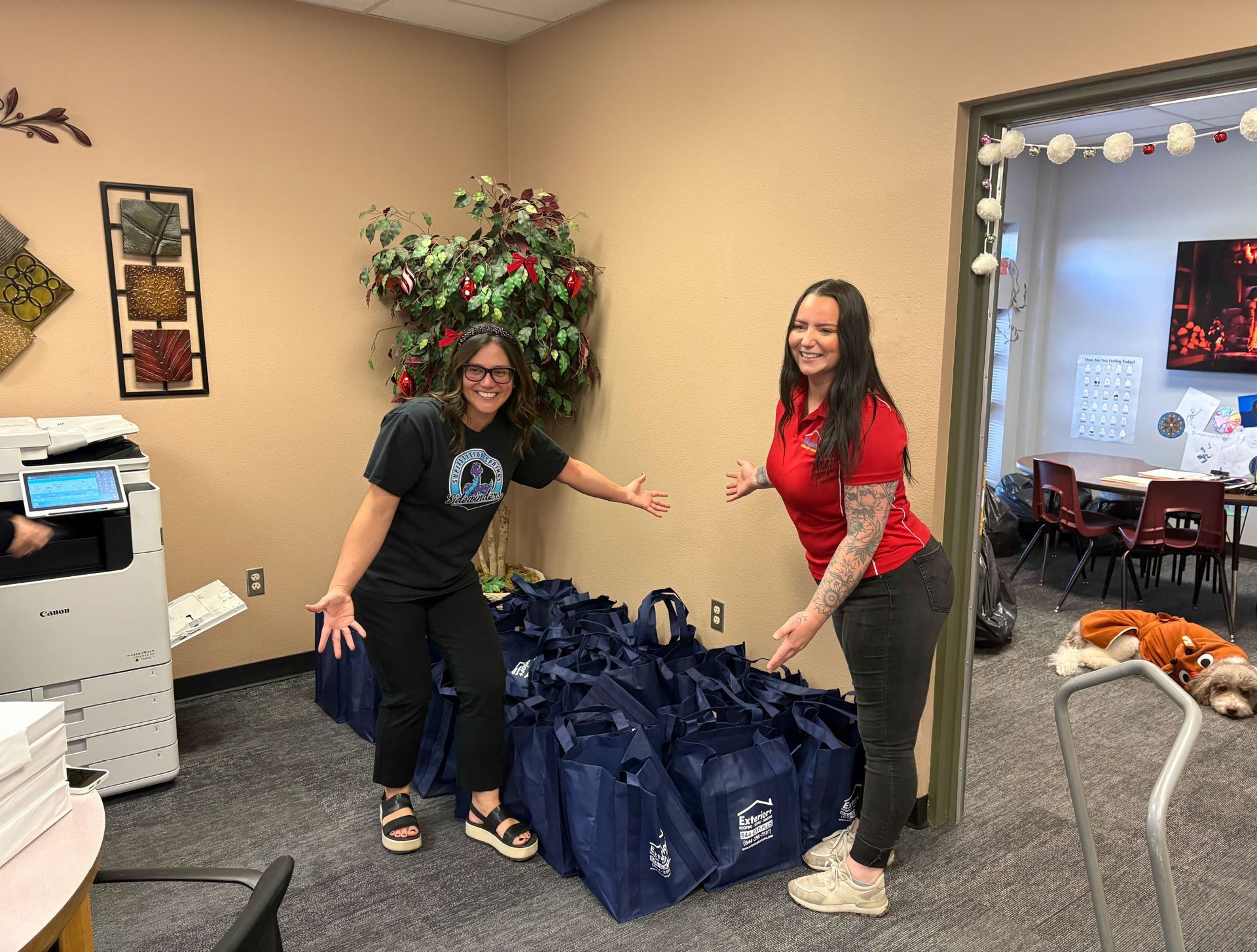 Two women pose beside a large stack of blue bags in an office. They are smiling, gesturing, and one has arm tattoos.