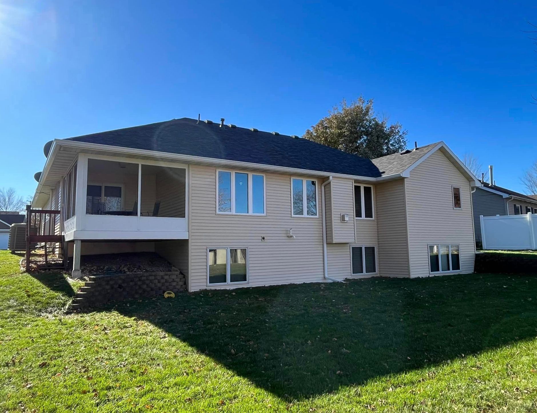 Back view of a two-story beige house with a screened porch and a dark blue roof, set in a grassy yard under a clear blue sky.