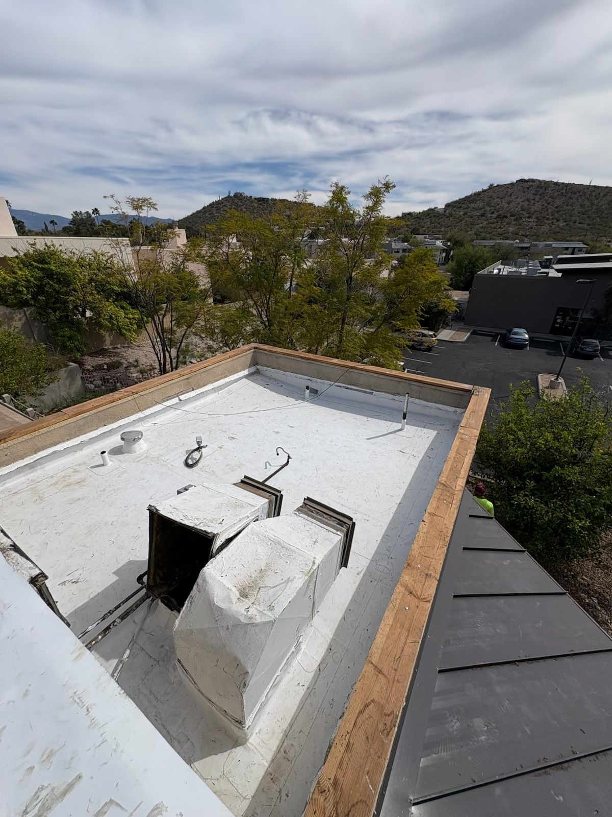 A high-angle view of a flat, white roof with two rectangular vents, framed by an exposed wooden edge against a desert landscape.