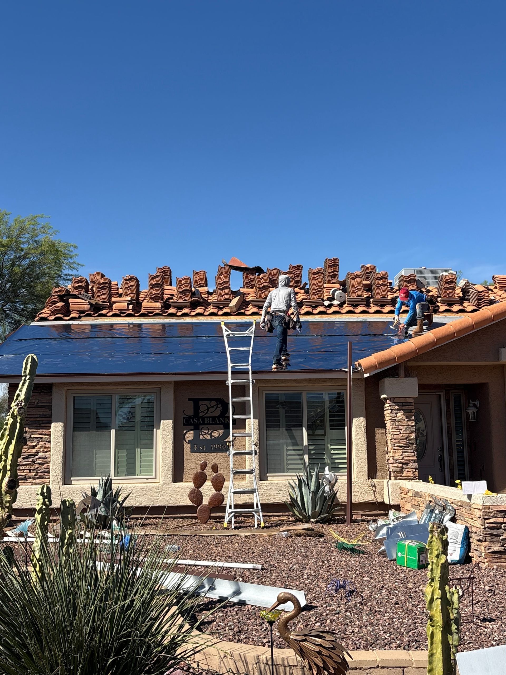 Two workers on a ladder and roof are performing repairs on a residential home with a partially removed tile roof.