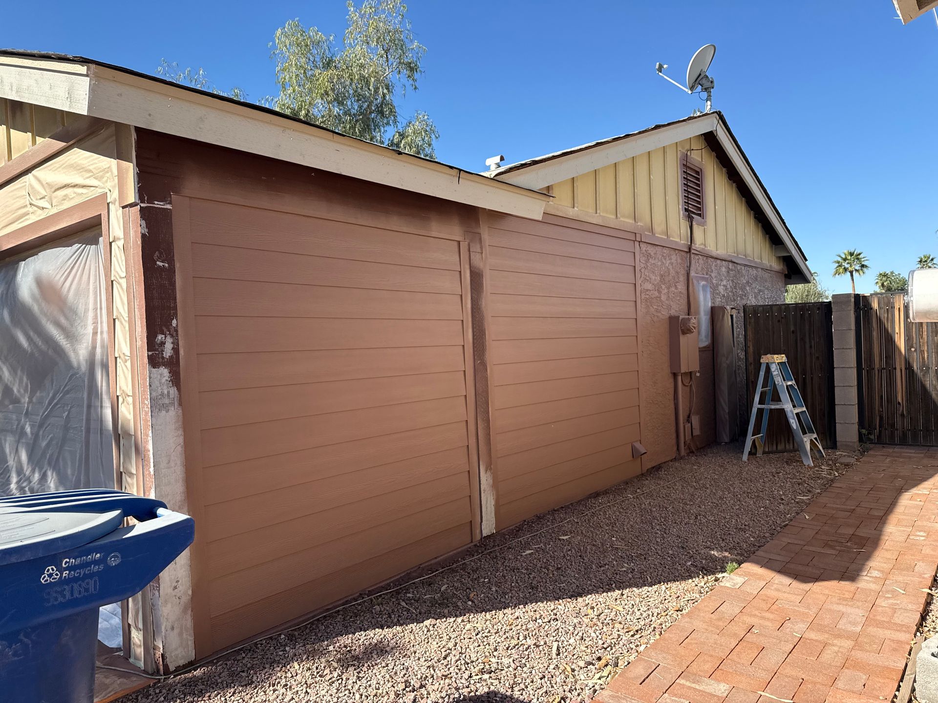 The exterior wall of a house undergoing renovations with tan siding, a blue trash bin, and a stepladder in the yard.