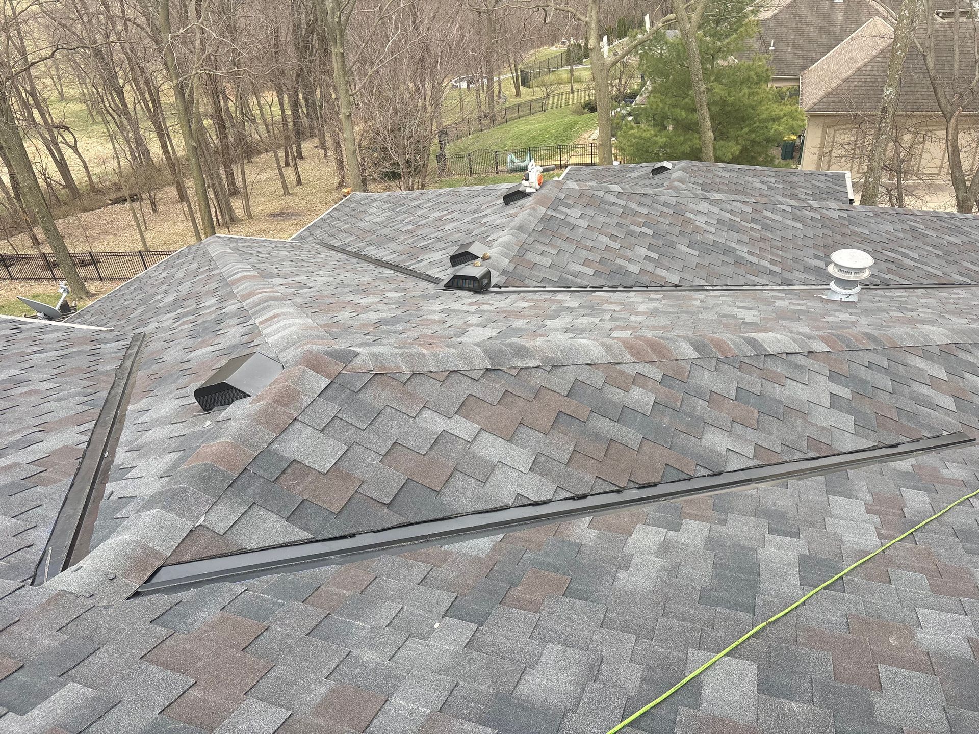 A residential shingled roof with several vents and metal flashing, viewed from above with trees in the background.