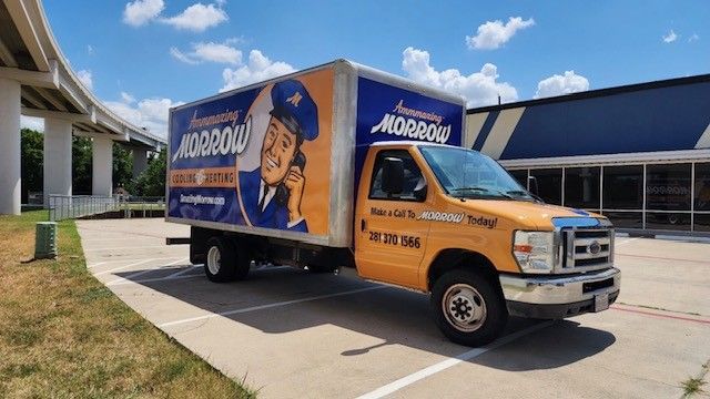 Yellow Morrow moving truck parked on pavement, blue sky, and a building in the background.