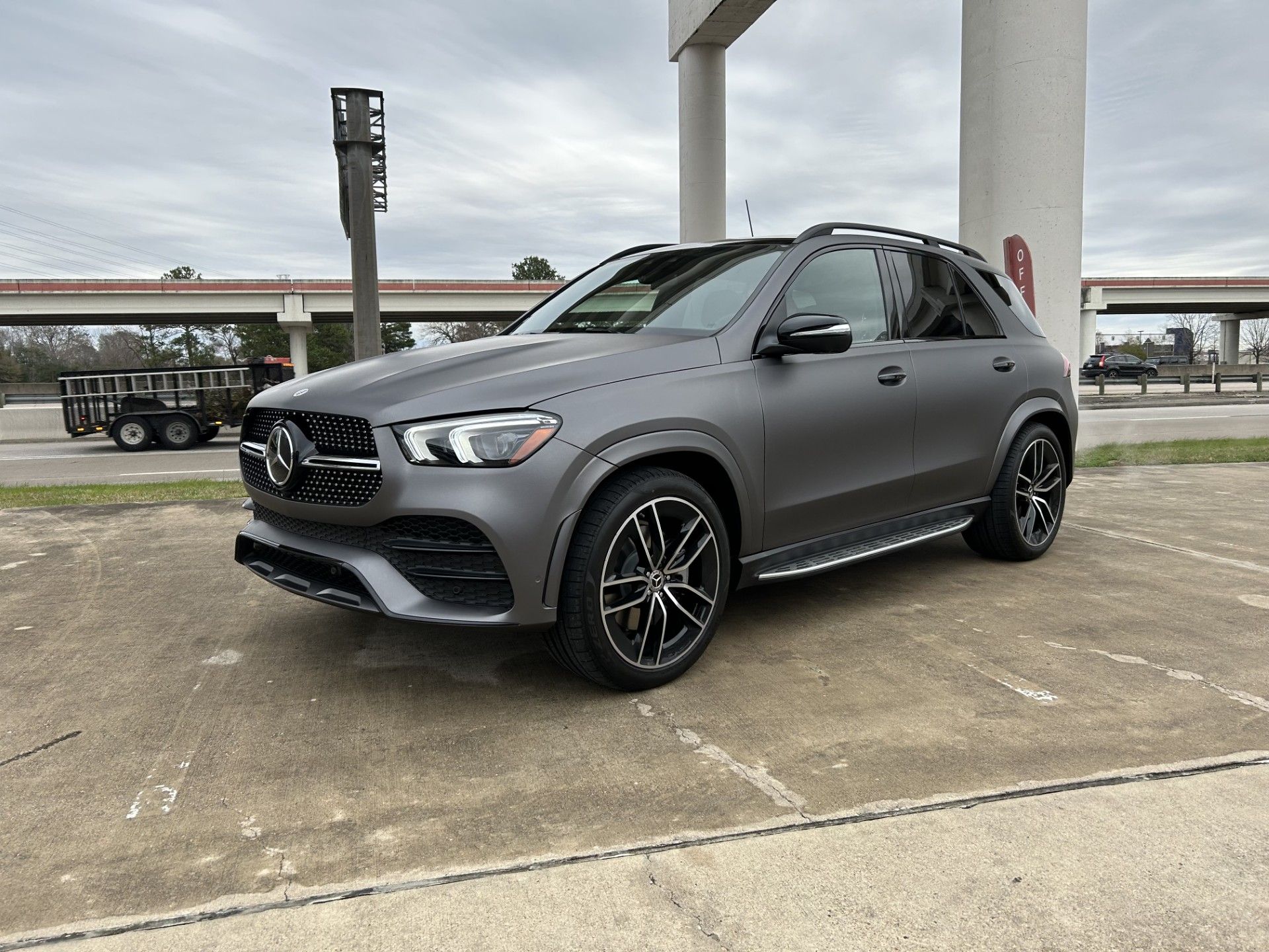 Gray Mercedes-Benz SUV parked on concrete near a pillar and highway overpass, overcast day.