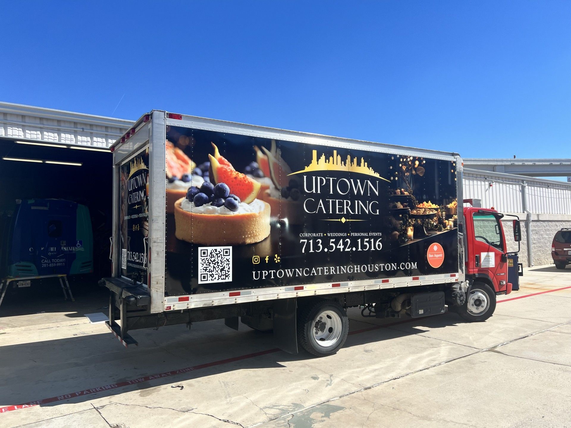 Uptown Catering truck with food graphic and contact info parked outside under a blue sky.