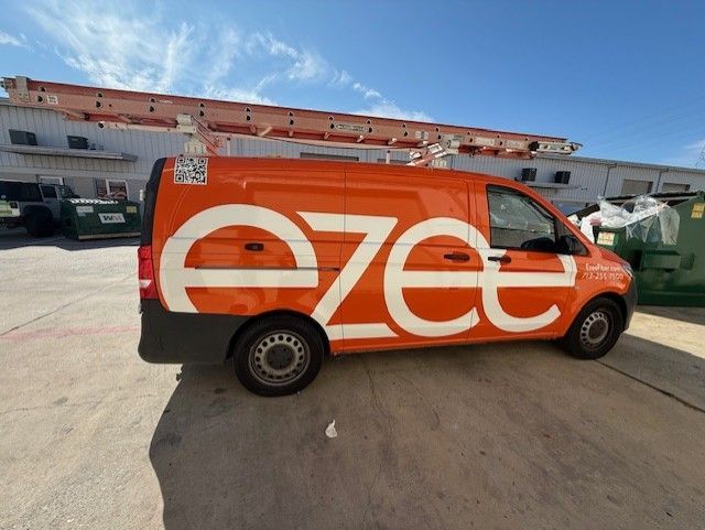 Orange ezee van with ladder on top, parked in front of a building on a sunny day.