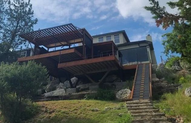 Two-story house with wooden deck, stairs, and a pergola, built on a grassy hillside. Cloudy sky.