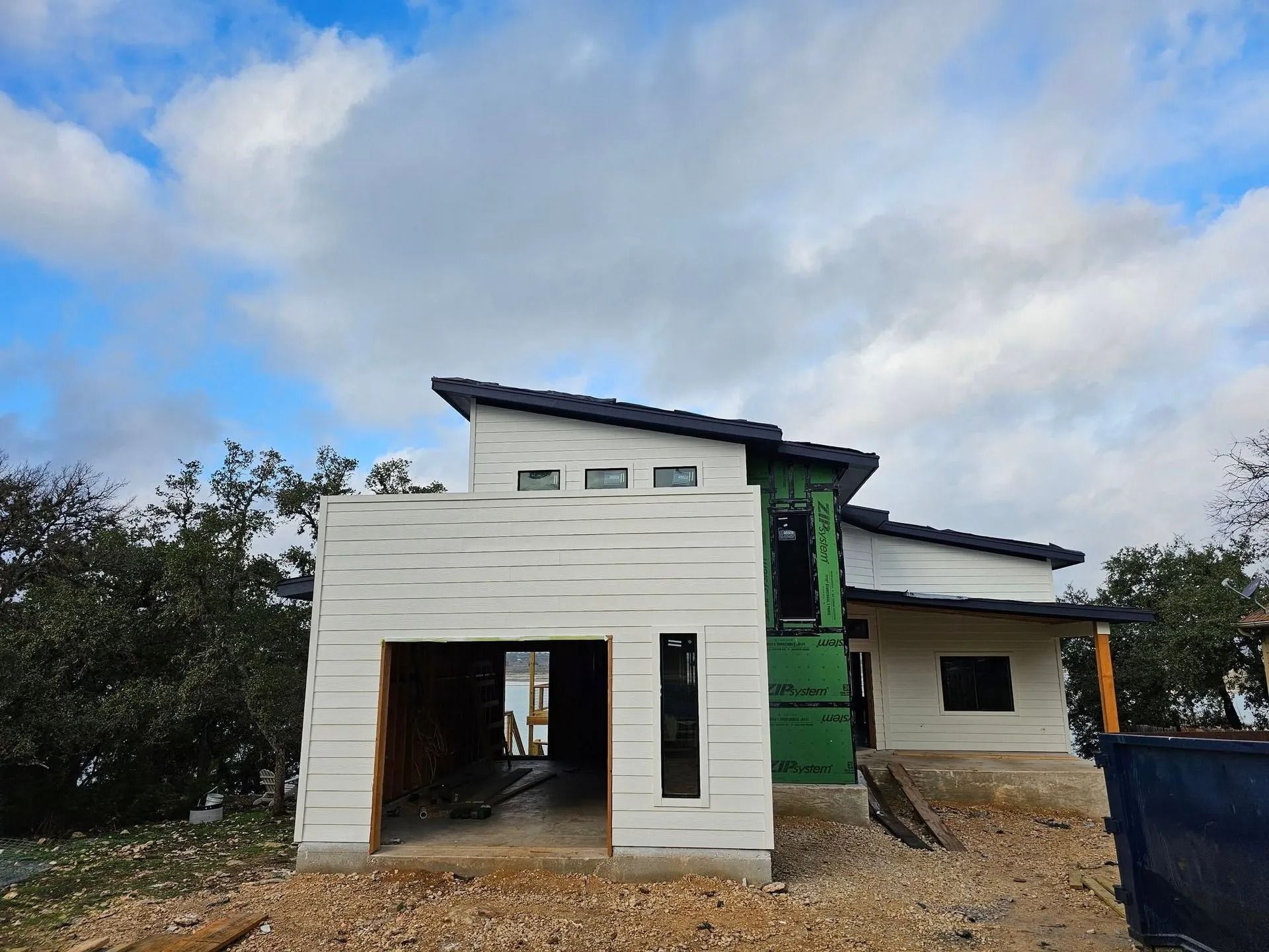 Modern house under construction with a garage, white siding, and a dark roof against a cloudy sky.