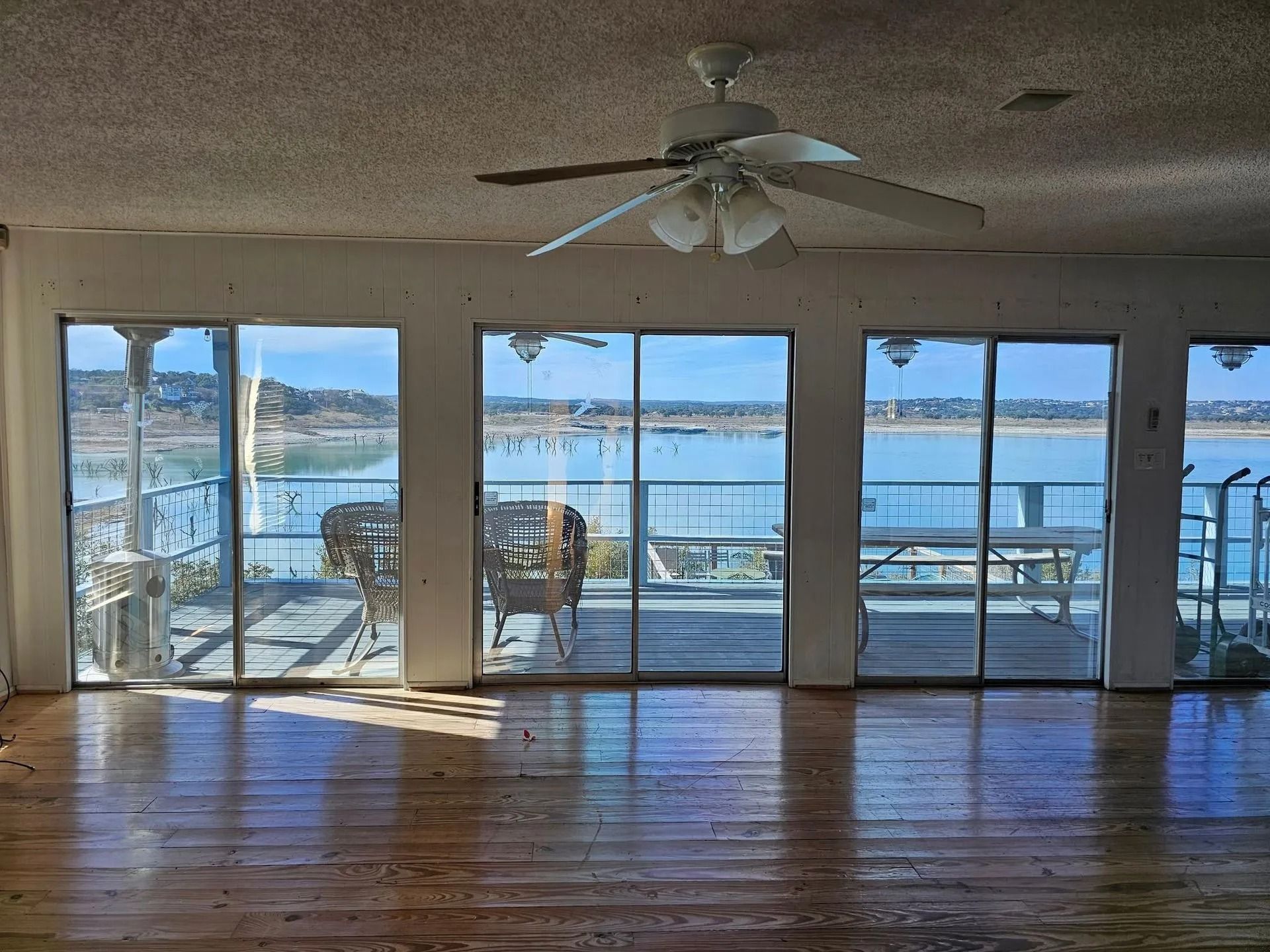 Interior view of a room with large windows overlooking a body of water and a patio; a ceiling fan is visible.