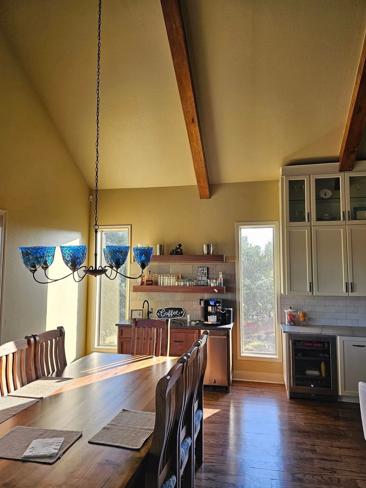 Dining room with long wooden table, blue chandelier, and kitchen with white cabinets.