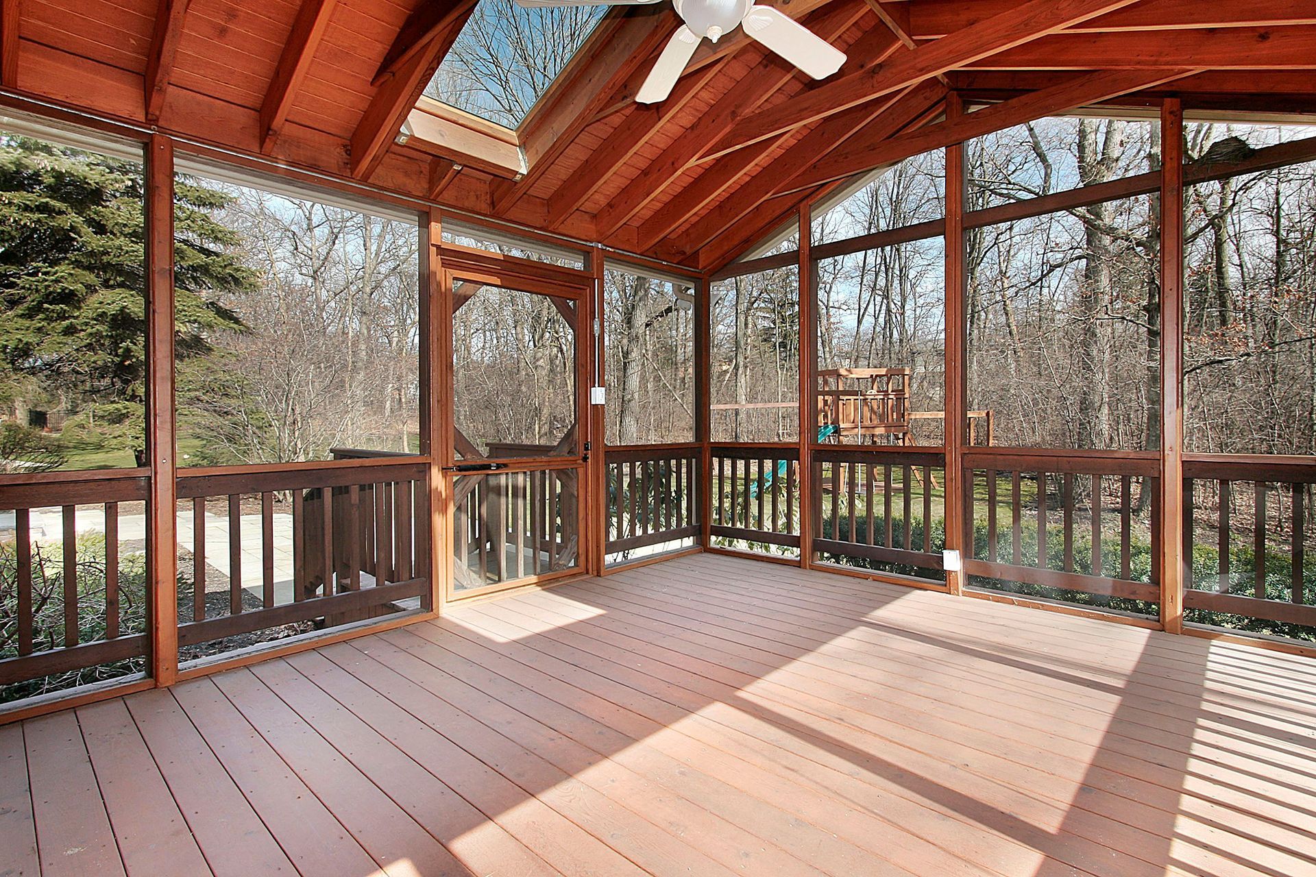 Screened porch with wooden deck, ceiling, and railing overlooking a wooded area.