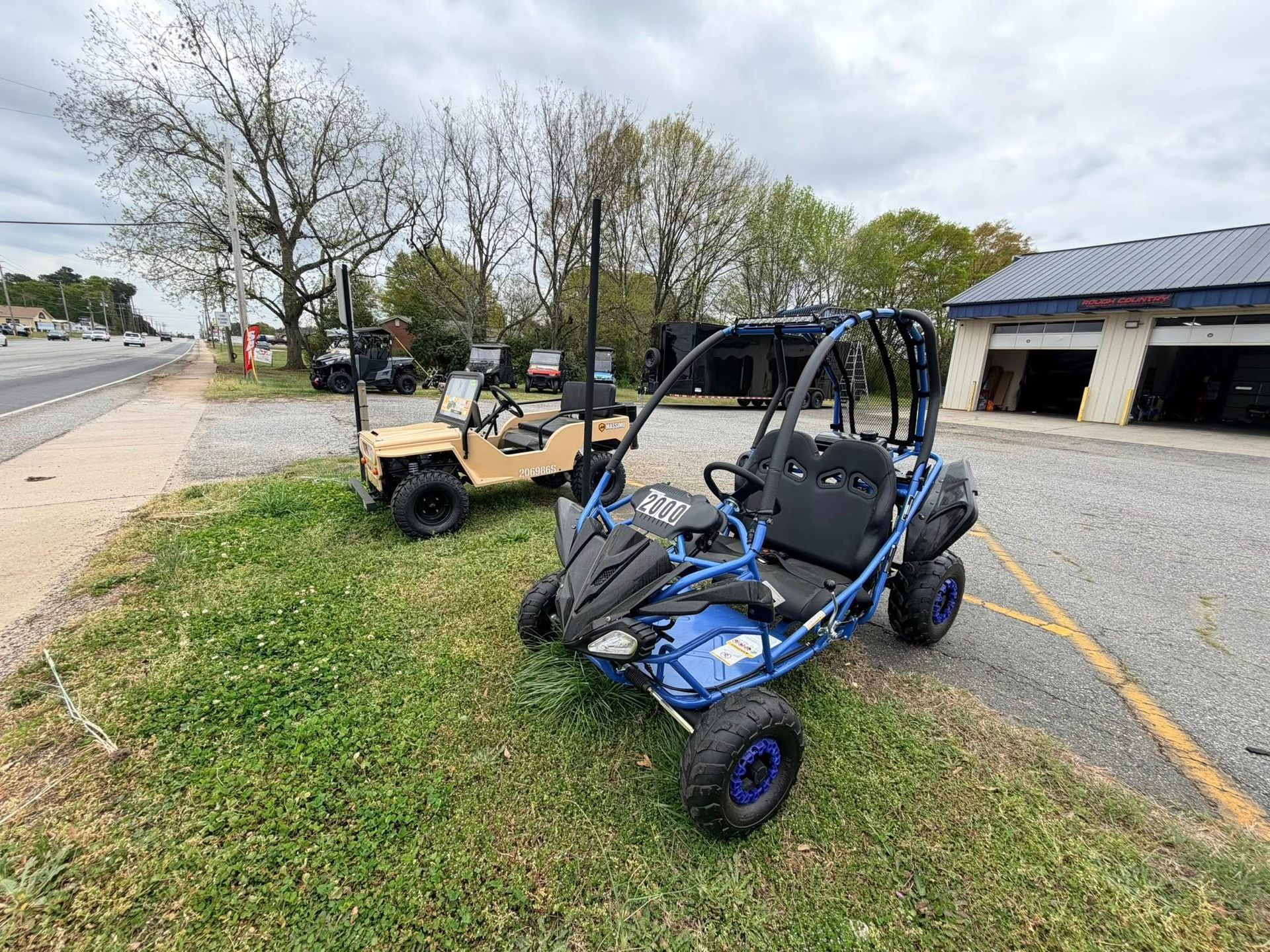 Blue off-road dune buggy parked on grass beside a road, with a tan ATV behind it and a building nearby