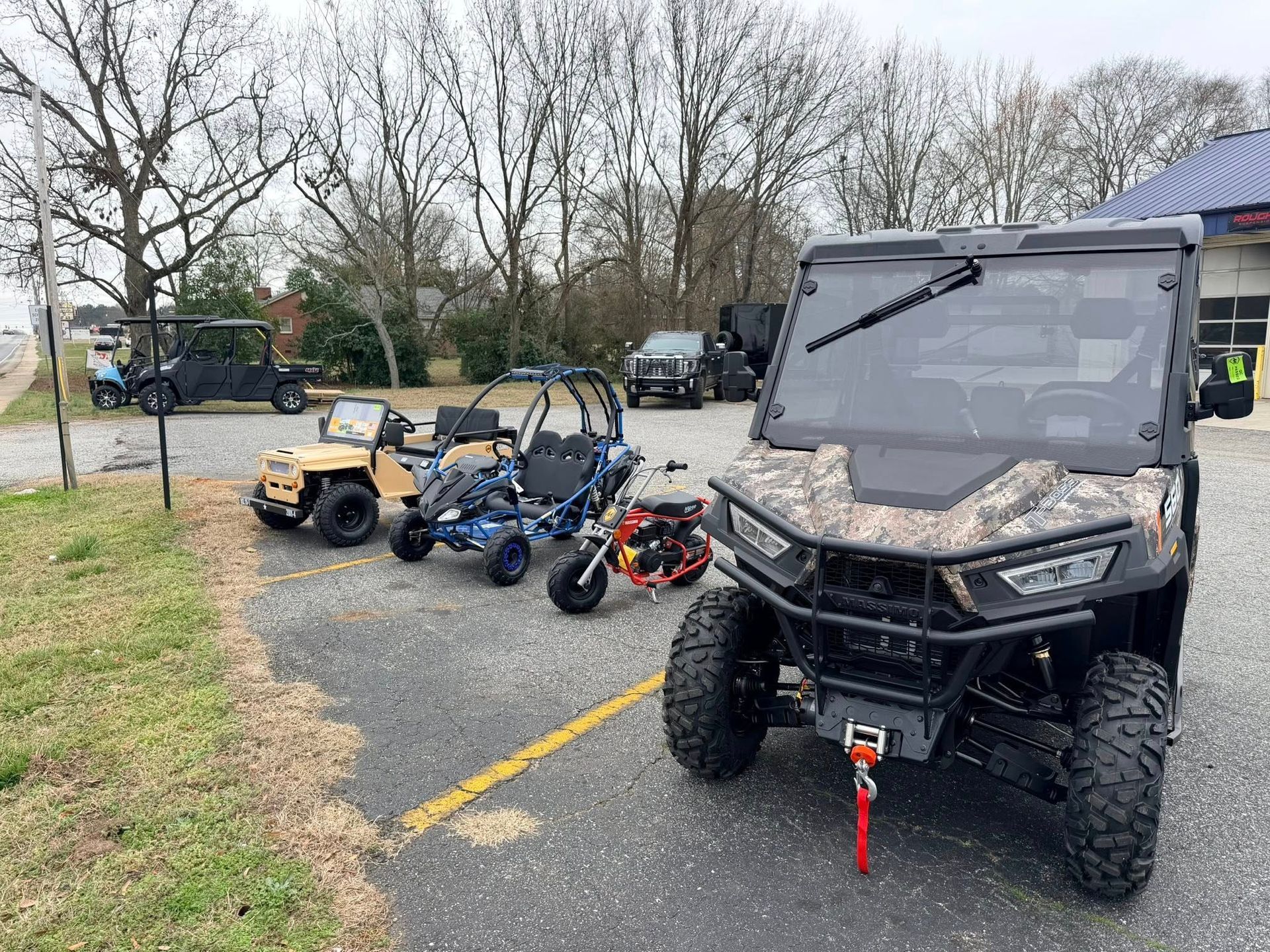 Off-road utility vehicles parked on a gravel lot near leafless trees, with a camo UTV in the foreground.