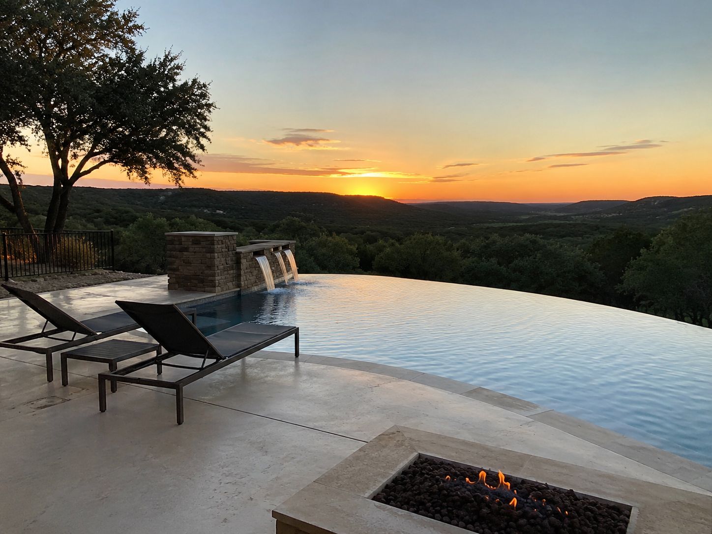 Infinity pool at sunset with lounge chairs, fire pit, and a distant green valley view