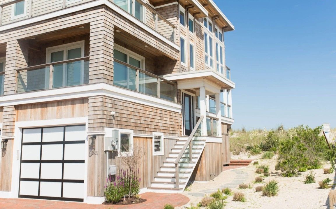 Two-story beach house with cedar siding, glass garage door, and wooden stairs leading to the entrance.
