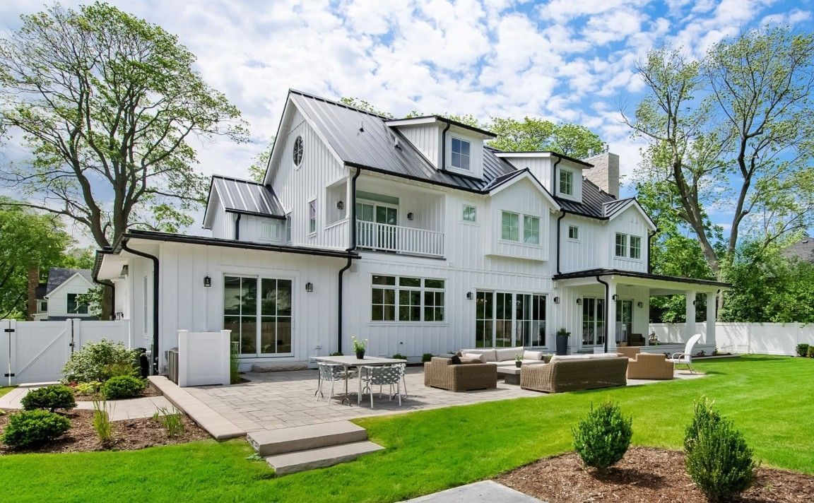 White farmhouse with black trim, a patio, and a green lawn under a blue sky.