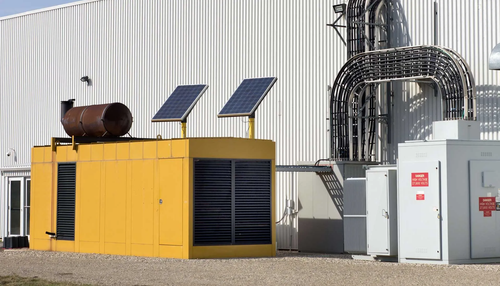 Yellow generator with solar panels, near a white building with electrical components and wiring.