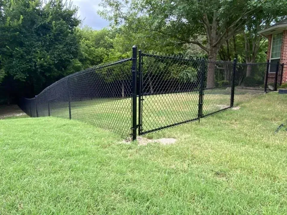 Black chain-link fence encloses a backyard, green grass, trees, and a house visible in the background.