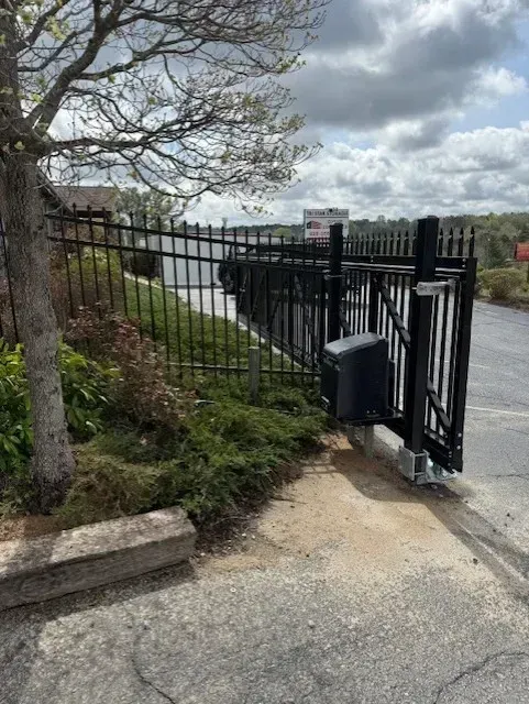 Black security gate, open, with fence, along a body of water, next to pavement, cloudy sky.