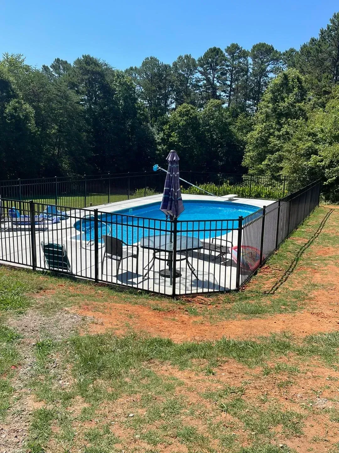 Fenced outdoor pool with umbrella, surrounded by trees. Clear blue water and sunny day.