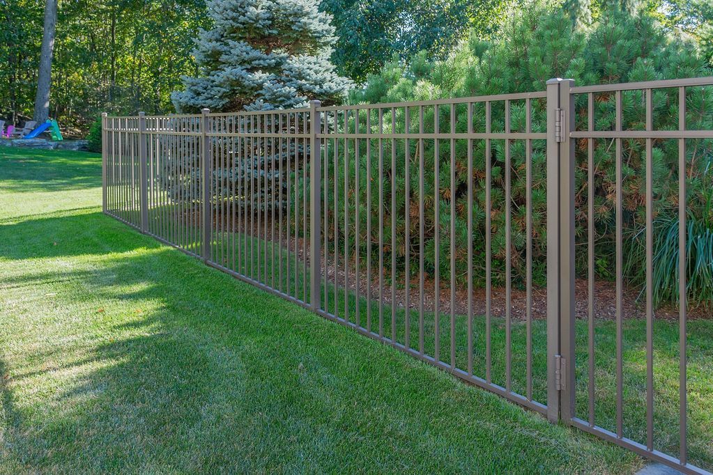 Brown metal fence in a grassy yard, with green trees and bushes in the background.