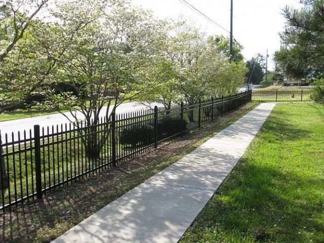 Black metal fence along a concrete sidewalk, with trees and green grass alongside the street.