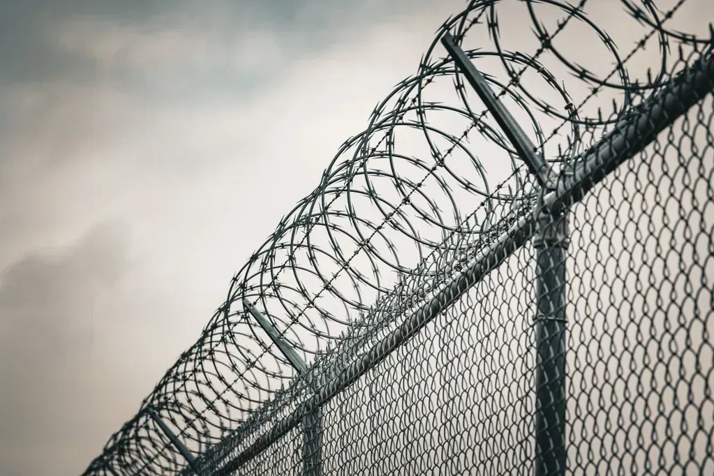 Barbed wire atop a chain-link fence against a cloudy sky.