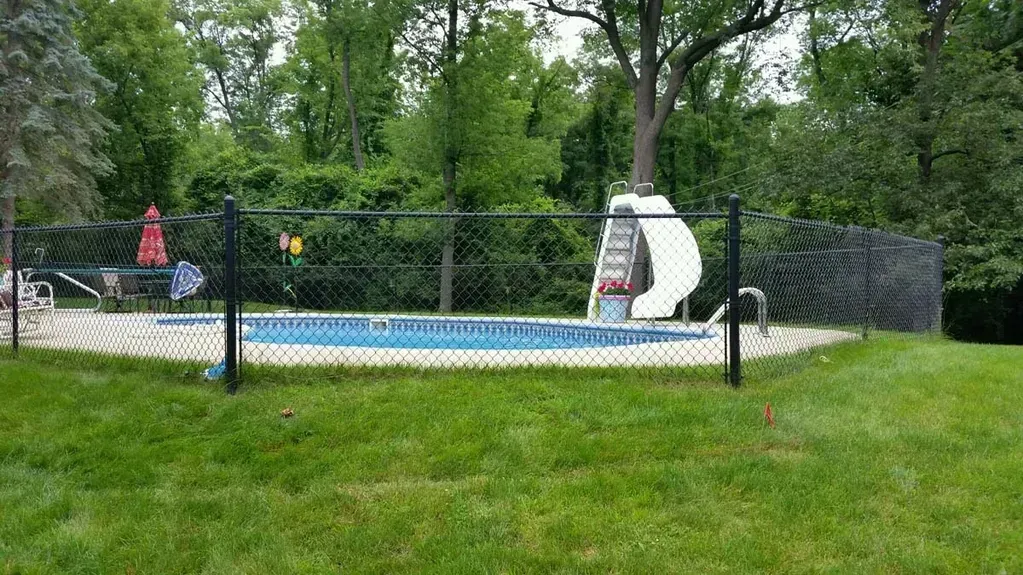 A backyard pool with a black chain-link fence, surrounded by green grass and trees.