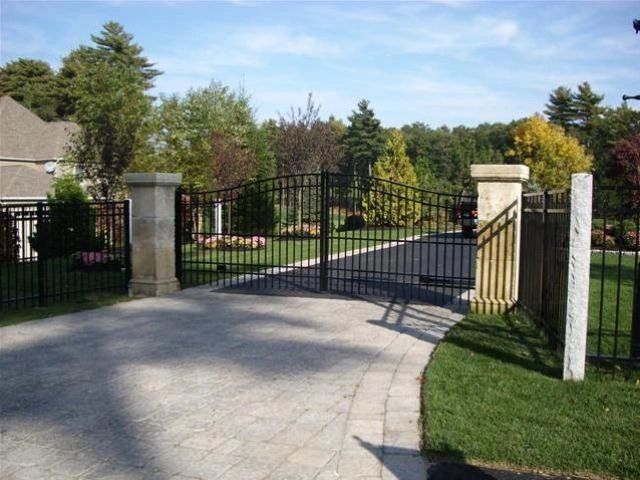A wrought iron gate on a stone driveway, flanked by stone columns and fencing, leads to a house.