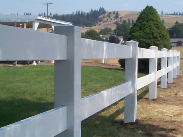 White ranch fence in a grassy yard with a hill in the background.