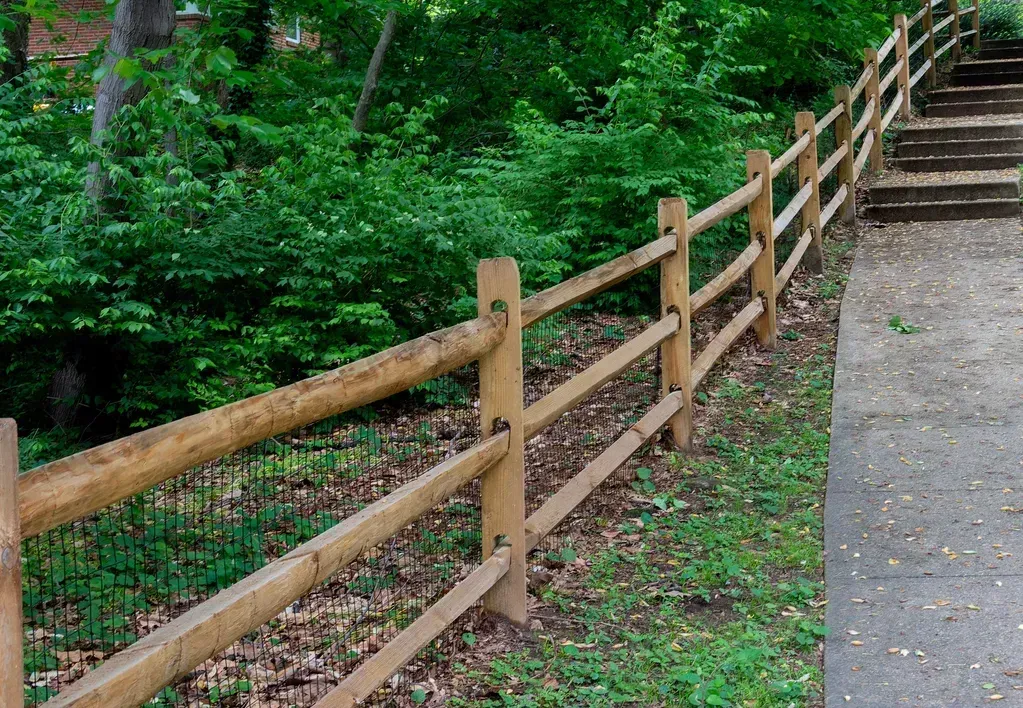 Wooden split-rail fence on a hill, alongside a paved path and steps, with lush green foliage.
