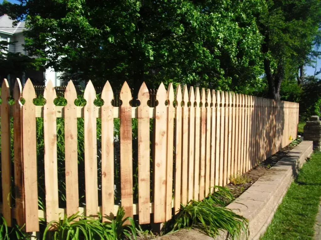 Wooden picket fence along a grassy verge, trees and a house in the background.