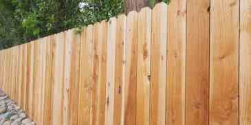 A wooden fence with a scalloped top, in a yard with foliage.