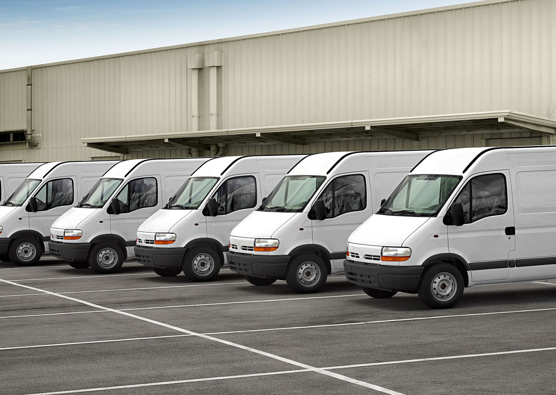 White delivery vans parked in front of a building on a paved lot under a blue sky.