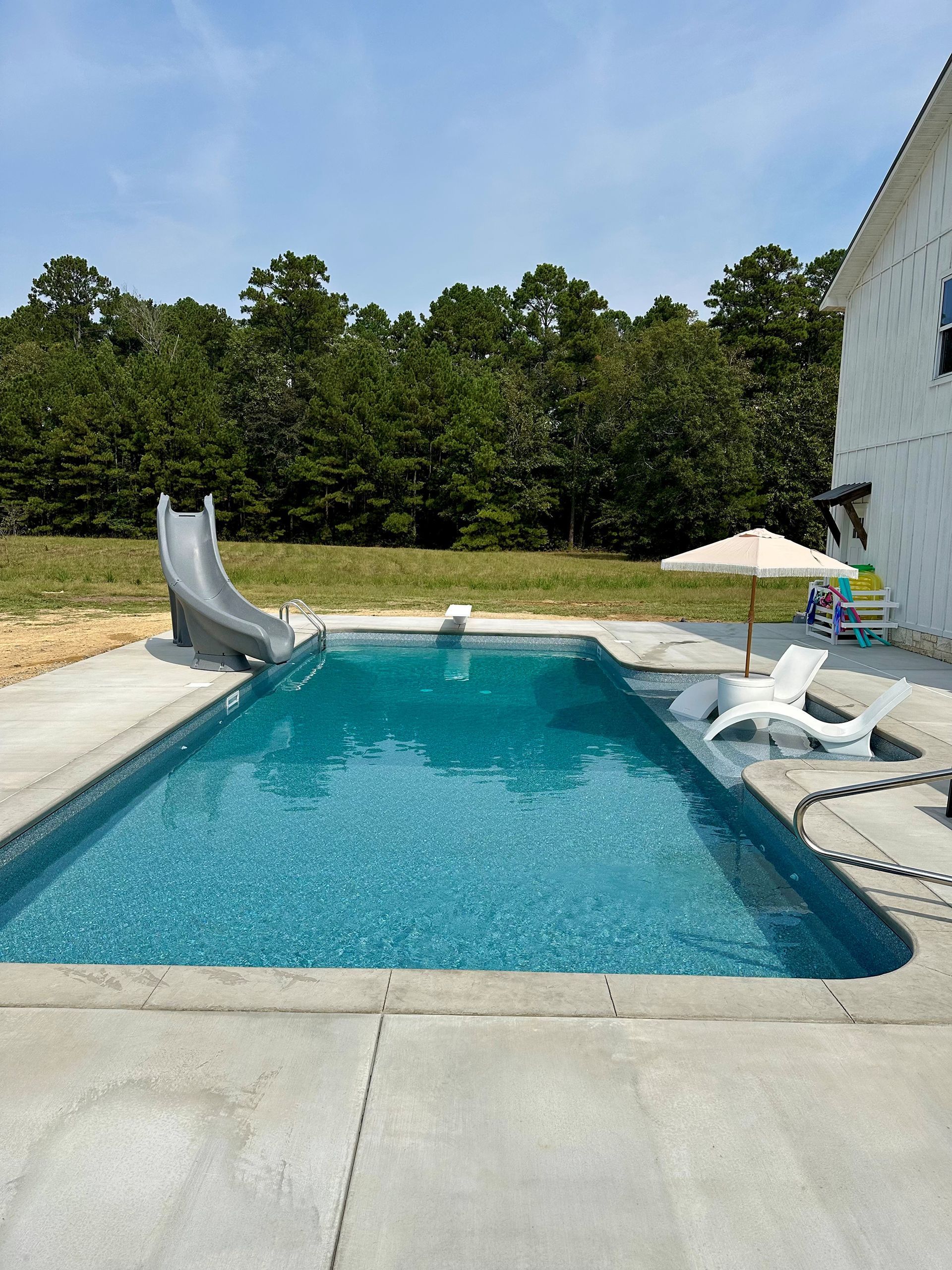 Pool with slide, umbrella, and lounge chairs. Green trees in background. Blue water. Sunny day.