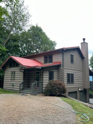 Two-story log cabin house with red roof and two garage doors; set on a hillside driveway with surrounding trees.