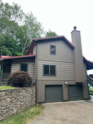 Log cabin-style house with gray siding, red roof, stone wall, and two-car garage. Overcast sky.
