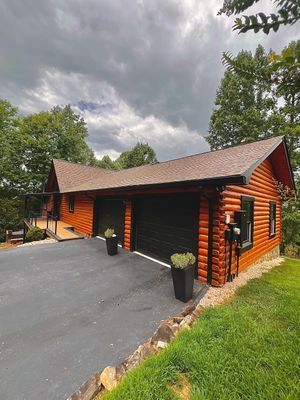 Log cabin with black garage doors, brown roof, and driveway on a grassy hill under a cloudy sky.